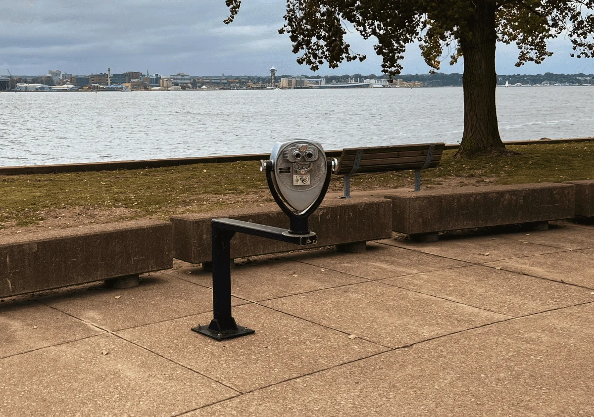 A Tower Optical ADA-compliant coin-operated binocular viewer installed by a waterfront walkway with benches, trees, and a city skyline in the background