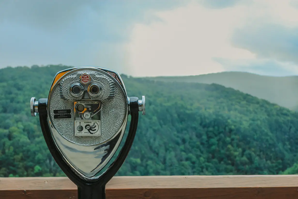 A Tower Optical coin-operated binocular viewer on a wooden platform overlooking a green mountain landscape under a cloudy sky