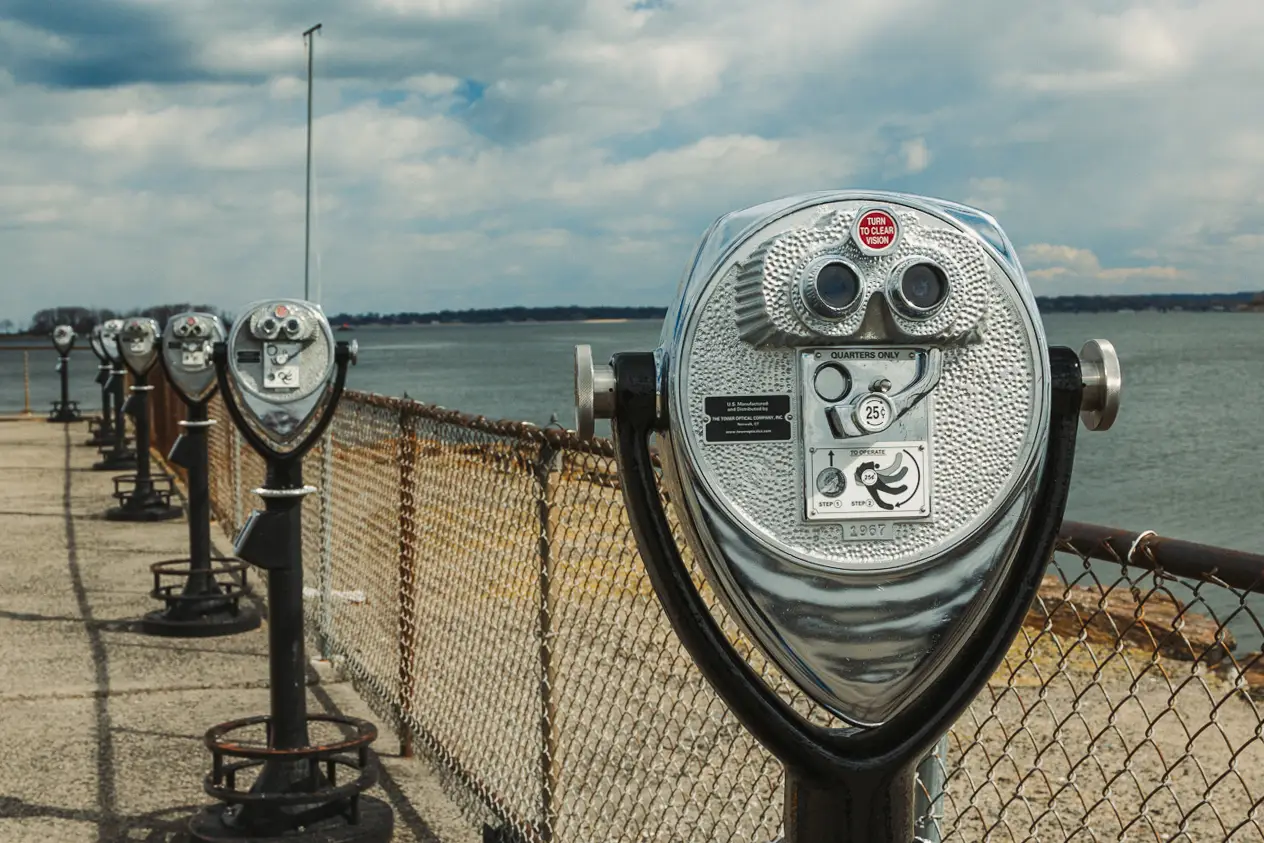 A row of Tower Optical coin-operated binocular viewers installed along a waterfront pier with a metal fence and ocean view in the background