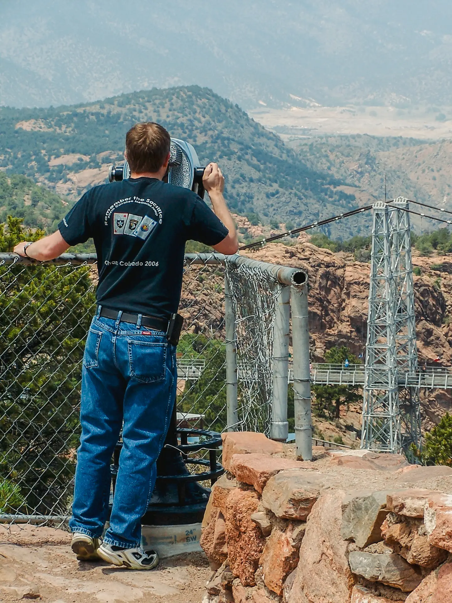 A man uses a Tower Optical coin-operated binocular viewer at a canyon overlook with a suspension bridge and mountains in the background