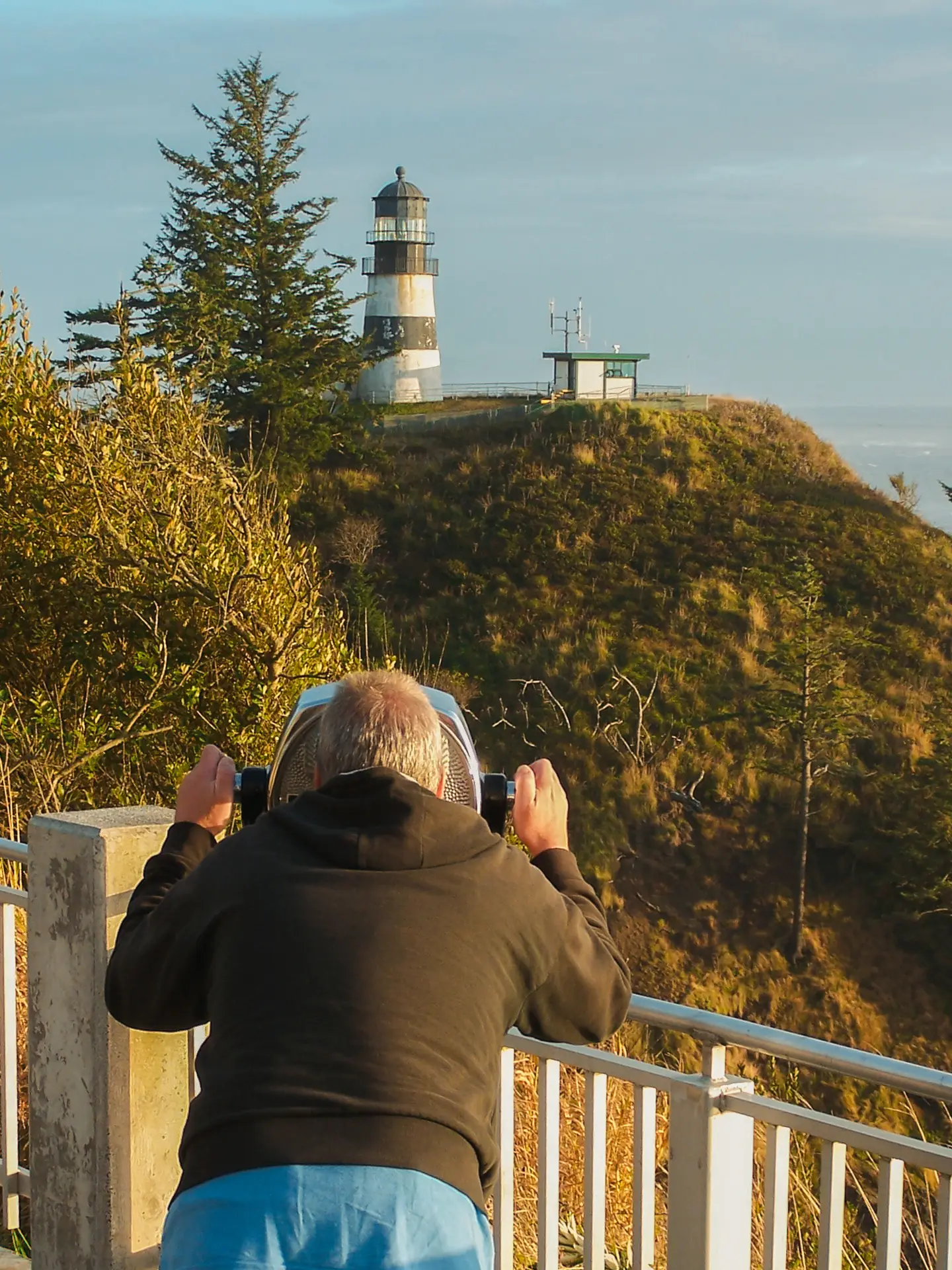 A person uses a Tower Optical coin-operated binocular viewer to look toward a lighthouse on a coastal cliff