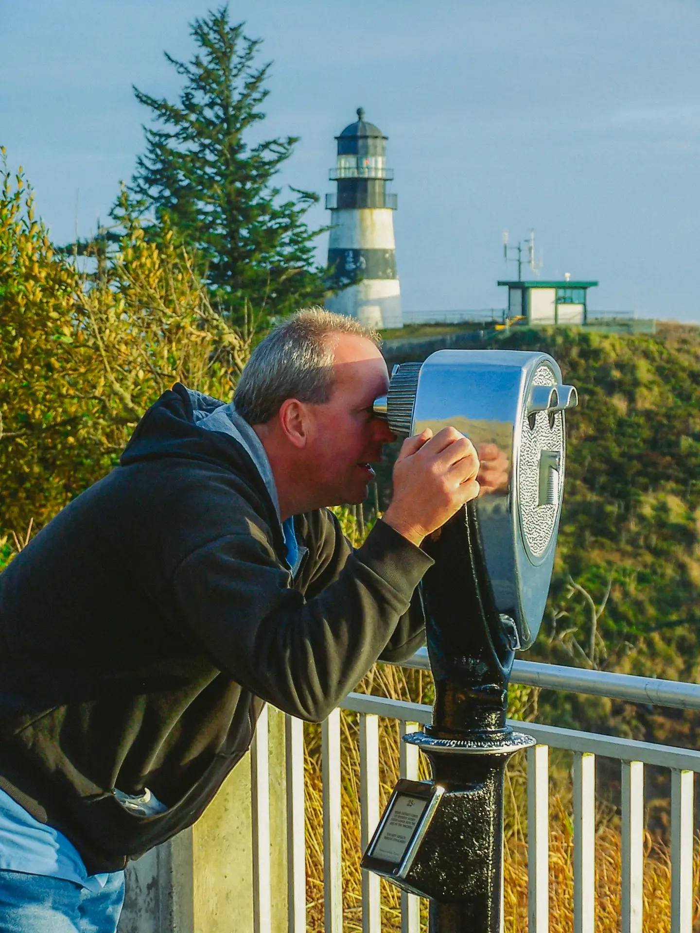 A man uses a Tower Optical coin-operated binocular viewer at a scenic overlook near a lighthouse