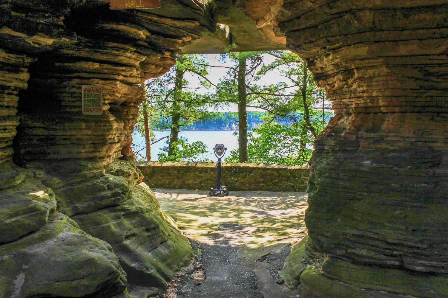 A Tower Optical coin-operated binocular viewer framed by a natural rock archway overlooking a scenic lake and forest