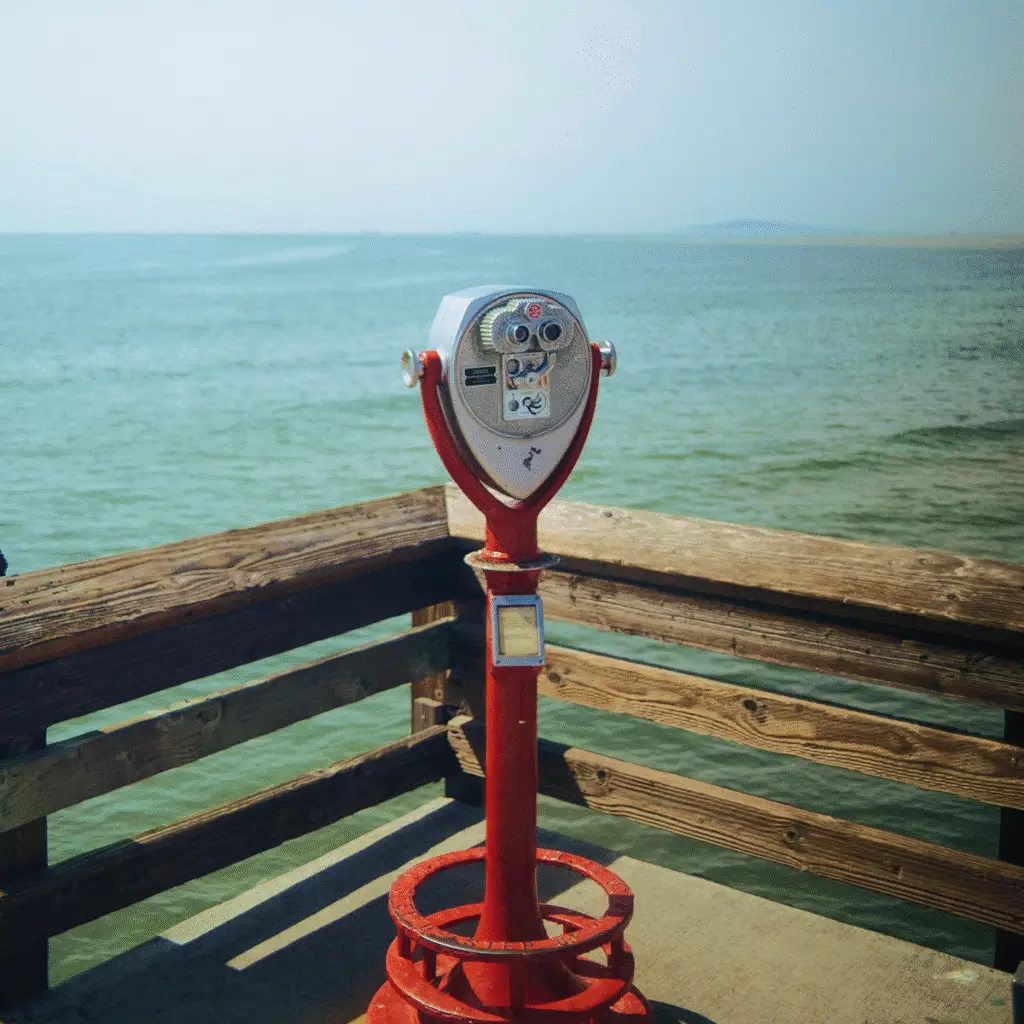 A red Tower Optical coin-operated binocular viewer mounted on a pier overlooking the ocean.