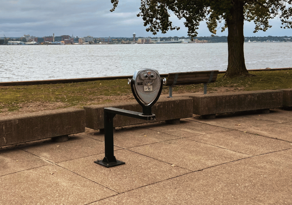 Tower Optical ADA-compliant coin-operated binocular viewer installed by a waterfront walkway with benches, trees, and city skyline in the background. Ski Resort