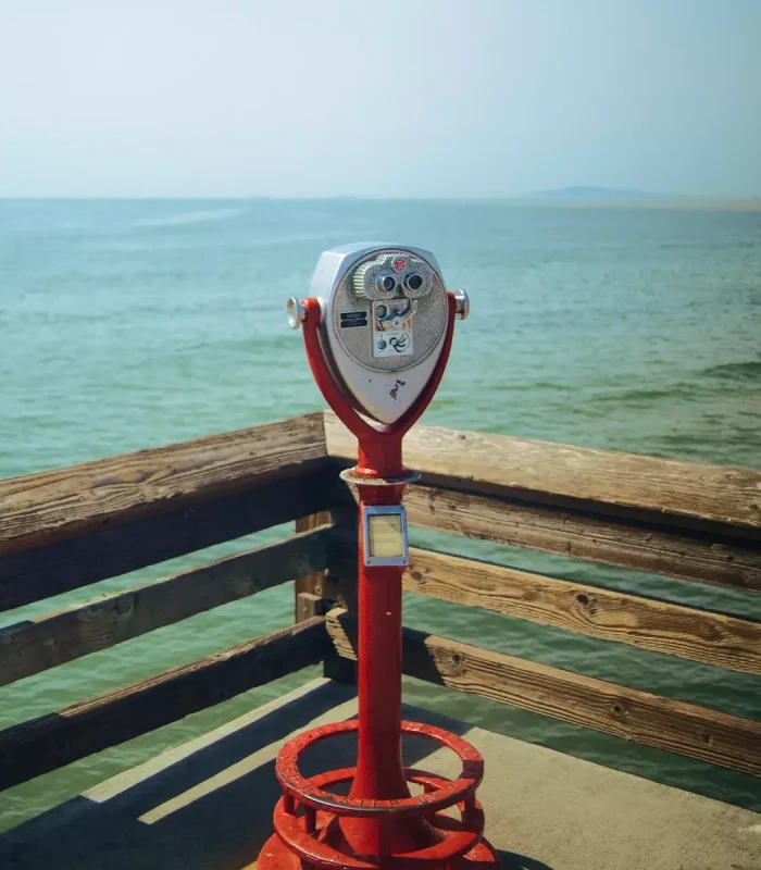 A red Tower Optical coin-operated binocular viewer mounted on a pier overlooking the ocean.